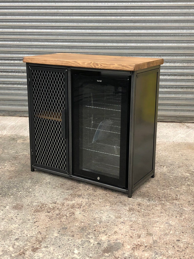 Pode incluir: Um m&oacute;vel retangular com estrutura de metal escuro, tampo de madeira natural e porta de malha met&aacute;lica preta &agrave; esquerda. O lado direito cont&eacute;m um refrigerador de bebidas embutido com porta de vidro, prateleiras de arame e acabamento preto.