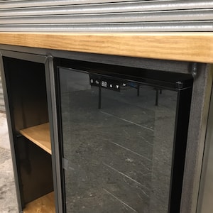 May include: A black metal wine cooler with a glass door and a wooden shelf. The cooler has a digital display with temperature controls. The cooler is sitting on a wooden countertop.