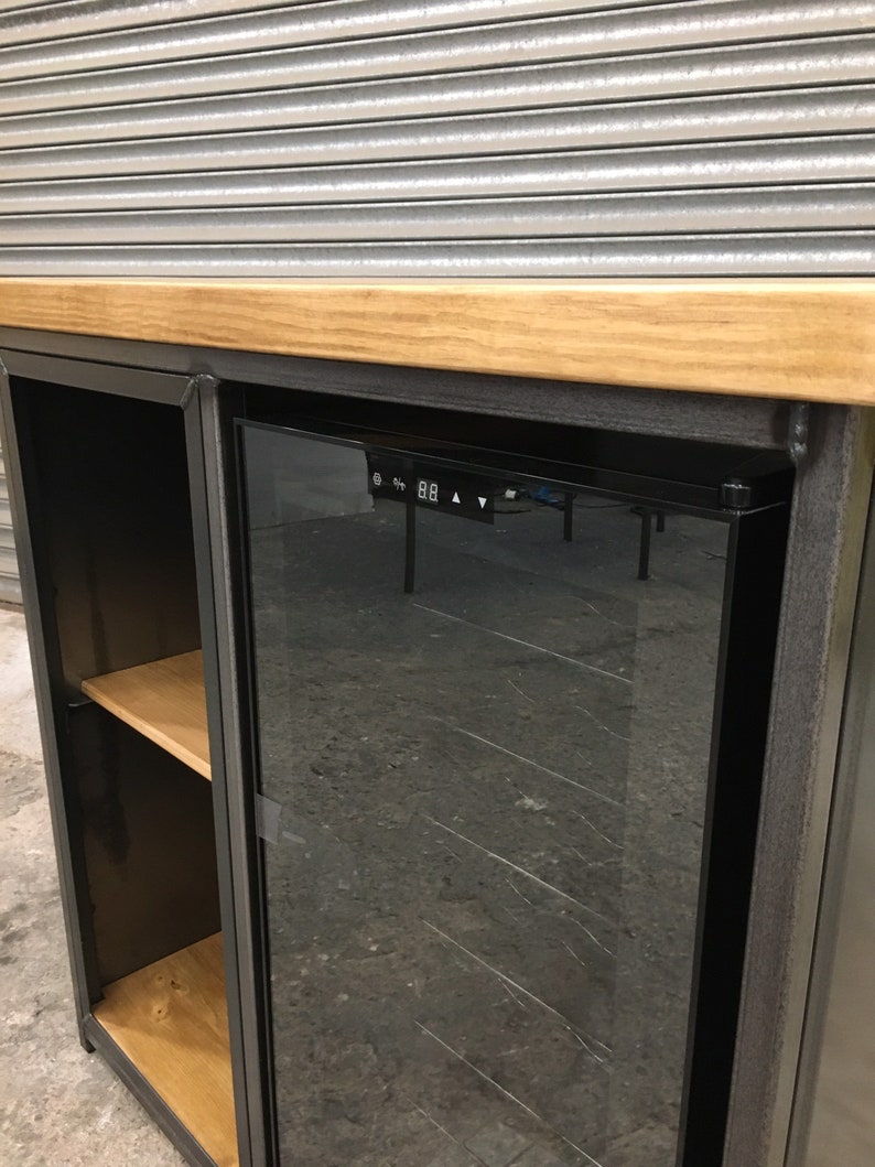 May include: A black metal wine cooler with a glass door and a wooden shelf. The cooler has a digital display with temperature controls. The cooler is sitting on a wooden countertop.