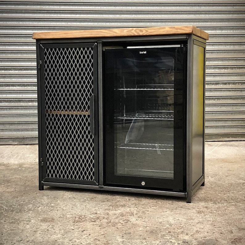 Pode incluir: Um m&oacute;vel retangular com estrutura de metal escuro, tampo de madeira natural e uma porta lateral com rede met&aacute;lica prateada em padr&atilde;o de losangos. Ao lado da porta, h&aacute; um refrigerador de bebidas preto com porta de vidro e prateleiras.