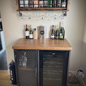 Pode incluir: Um m&oacute;vel bar de madeira com porta de malha met&aacute;lica e um refrigerador de bebidas com porta de vidro, com uma superf&iacute;cie de madeira que sustenta garrafas. Acima, uma prateleira de parede de madeira cont&eacute;m garrafas e ta&ccedil;as de vinho penduradas.