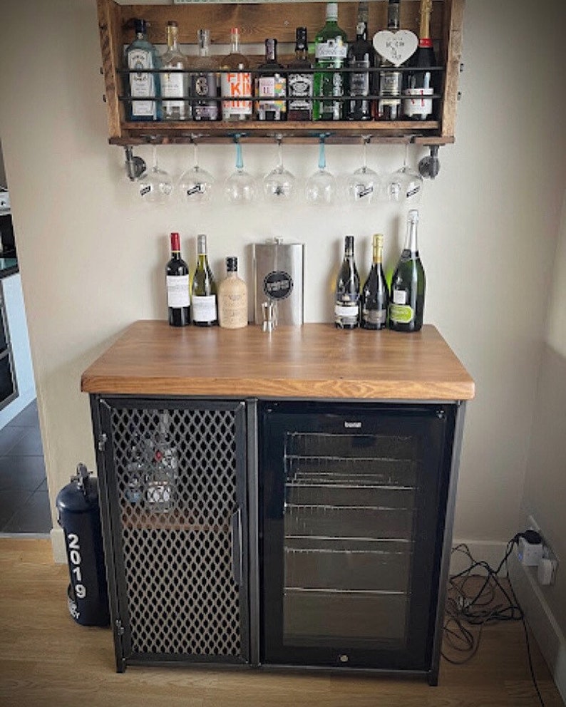 Pode incluir: Um m&oacute;vel bar de madeira com porta de malha met&aacute;lica e um refrigerador de bebidas com porta de vidro, com uma superf&iacute;cie de madeira que sustenta garrafas. Acima, uma prateleira de parede de madeira cont&eacute;m garrafas e ta&ccedil;as de vinho penduradas.