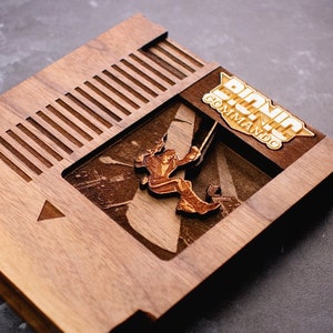 May include: A wooden replica of a classic Nintendo Entertainment System game cartridge featuring the title "Bionic Commando" in gold lettering. The cartridge is made of walnut wood and has a cutout of the game's cover art.
