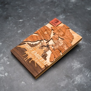 Könnte beinhalten: Ein hölzerner Spielkoffer für das Nintendo Switch-Spiel Fire Emblem: Three Houses. Der Koffer ist aus hellem und dunklem Holz gefertigt und zeigt ein 3D-Lasercut-Design von Charakteren aus dem Spiel.