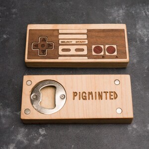 Pode incluir: Dois abridores de garrafas de madeira. O superior tem a forma de um controlador de videogame clássico. O inferior é retangular com um abridor de garrafas e a palavra "PIGMINTED" gravada. Em uma superfície cinza escuro.