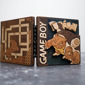May include: A wooden Game Boy-style box with a hinged design. One side features a maze pattern, while the other displays a Pokémon character and the word "Pokémon." The box is made of various wood tones and has the words "GAME BOY" on the side.