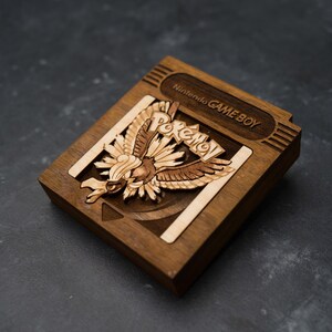 May include: Wooden Game Boy cartridge case featuring a laser-cut design. The case is brown with a light-colored bird and the word "Pokémon" in the center. The top reads "Nintendo GAME BOY".