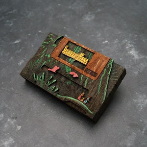 Puede incluir: Una caja de madera con un diseño tallado de una escena de bosque. La caja está pintada en tonos de verde, marrón y amarillo. Hay pequeñas figuras de personas talladas en la escena.