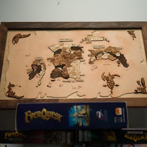 May include: A wooden map of a fantasy world, framed in dark wood. The map features detailed landmasses and is labeled with place names. Decorative elements are placed around the map. The EverQuest game box is visible below the map.