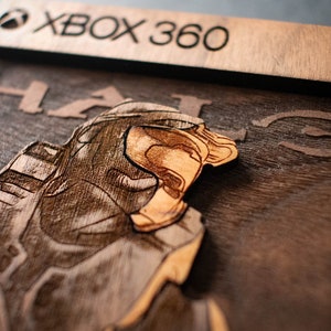 Puede incluir: Una placa de madera con una imagen grabada con láser de un Jefe Maestro de la serie de videojuegos Halo. La placa es marrón y presenta el texto "XBOX 360" y "HALO" en una fuente estilizada.