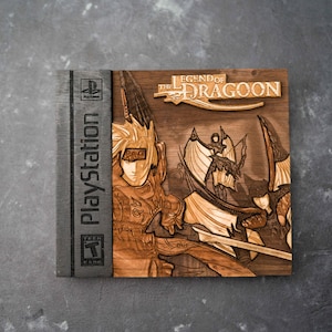 May include: A wooden laser-cut replica of the PlayStation game case for The Legend of Dragoon. The case features a brown wood background with a detailed image of the game's main character, a young man with blonde hair and a blue bandana, holding a sword. The game title is printed in white on a black banner.