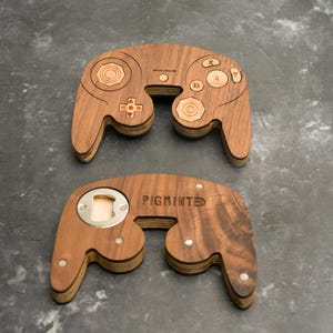 May include: Two wooden bottle openers shaped like video game controllers. The top one has controller buttons etched into the wood. The bottom one has a bottle opener and the word "PIGMINTED" etched into the wood.