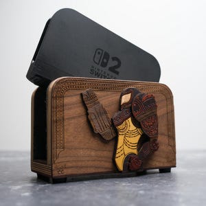 以下が含まれることがあります： 黄色と茶色のスーツを着たキャラクターが特徴の、木製の Nintendo Switch ドック。ドックは茶色で、上部は黒色、Nintendo Switch のロゴが見えます。