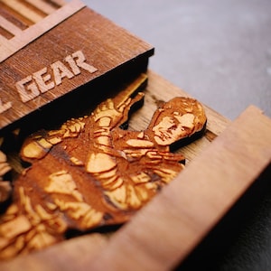 Puede incluir: Una caja de madera con las palabras "METAL GEAR" grabadas en la parte superior. La caja está abierta y en su interior hay una imagen de madera cortada con láser de un personaje de la serie de videojuegos Metal Gear.