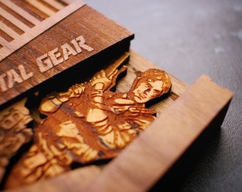 May include: A wooden box with the words "METAL GEAR" engraved on the top. The box is open and inside there is a laser-cut wooden image of a character from the Metal Gear video game series.