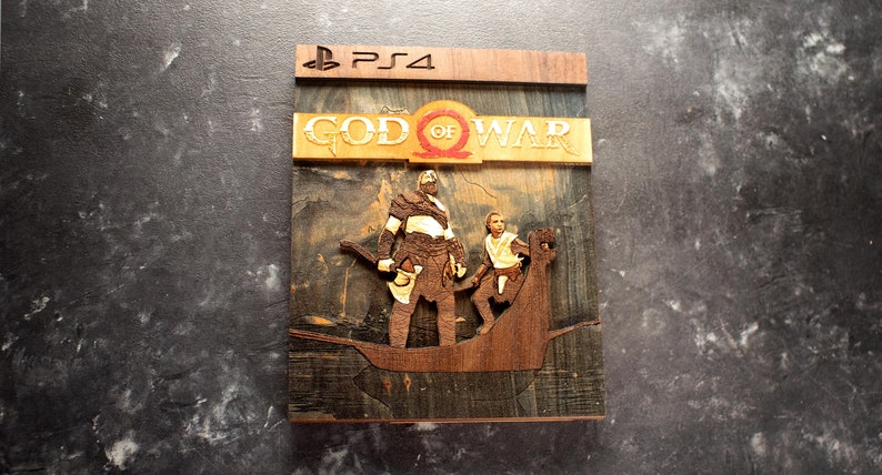 Pode incluir: Uma caixa de jogo de madeira para o jogo God of War para PlayStation 4. A caixa apresenta uma imagem queimada em madeira de Kratos e Atreus em p&eacute; em um barco. O t&iacute;tulo "God of War" est&aacute; escrito em letras douradas na parte superior da caixa.