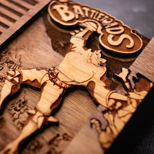 May include: A wooden box with a carved image of a character from the video game Battletoads. The character is holding a large, round object above its head. The box is made of dark wood and has a light brown finish. The box is labeled "Battletoads" in a stylized font.
