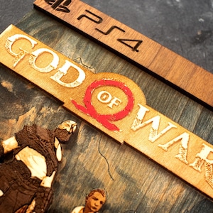 Pode incluir: Uma capa de jogo de madeira para o jogo God of War para PlayStation 4. A capa apresenta uma imagem 3D de Kratos e Atreus do jogo, juntamente com o t&iacute;tulo e o logotipo do jogo.
