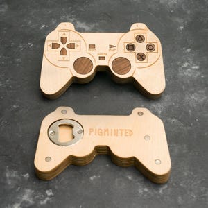 Pode incluir: Dois abridores de garrafa de madeira em forma de comando de jogo. O superior tem botões gravados e um centro cor de noz. O inferior tem um abridor de garrafa e a palavra "PIGMINTED" gravada. Estão sobre uma superfície cinza escuro.