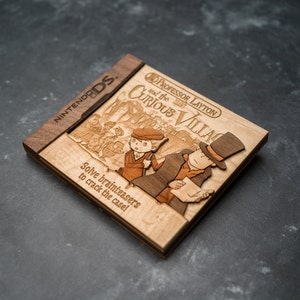 Puede incluir: Una caja de juego de Nintendo DS de madera para Professor Layton y el Pueblo Misterioso. La caja está hecha de madera clara con un borde de madera oscura. La parte delantera de la caja presenta una imagen grabada con láser del profesor Layton y Luke Triton. El texto "¡Resuelve acertijos para resolver el caso!" también está grabado en la caja.