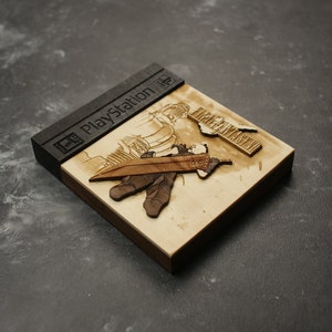 K&ouml;nnte beinhalten: Ein h&ouml;lzerner PlayStation-Spielekoffer mit einem Laser-Cut-Design eines Charakters aus Final Fantasy VII. Der Koffer ist schwarz und hat das PlayStation-Logo auf der Vorderseite. Der Charakter h&auml;lt ein Schwert und steht vor einem Geb&auml;ude.