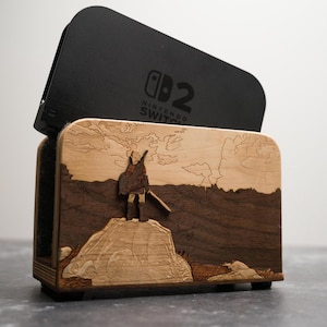 Op de afbeelding: Houten Nintendo Switch-dock met een lasergeëtst landschapsontwerp. De dock heeft een zwarte bovenkant met het Nintendo Switch-logo. Het voorpaneel toont een gedetailleerde scène met een figuur op een rots, in bruin- en beigetinten.
