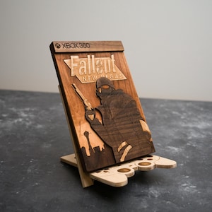 Puede incluir: Soporte de consola de juegos de madera con la carátula del juego Fallout: New Vegas. El soporte está hecho de madera clara y oscura, con el logotipo de Xbox 360 en la parte superior. El soporte tiene una base en forma de mando.