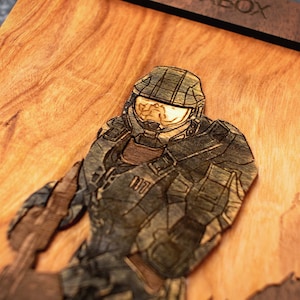 May include: A wooden wall art piece featuring a 3D laser-cut design of a Master Chief from the Halo video game series. The artwork is made of wood with a natural finish and has a detailed design.