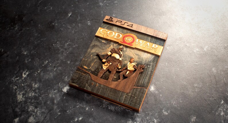 Pode incluir: Uma r&eacute;plica de madeira da capa do jogo God of War para PS4. A capa apresenta uma escultura detalhada de Kratos e Atreus em um barco, com o t&iacute;tulo do jogo "God of War" em letras douradas.