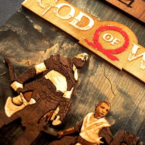 Pode incluir: Uma escultura 3D de madeira da capa do jogo God of War. A escultura apresenta Kratos e Atreus em p&eacute; em frente a uma cordilheira. O t&iacute;tulo "God of War" &eacute; exibido em uma fonte de madeira em relevo.