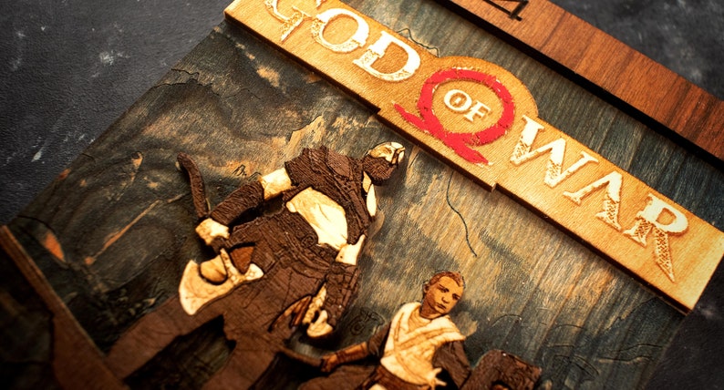 Pode incluir: Uma escultura 3D de madeira da capa do jogo God of War. A escultura apresenta Kratos e Atreus em p&eacute; em frente a uma cordilheira. O t&iacute;tulo "God of War" &eacute; exibido em uma fonte de madeira em relevo.