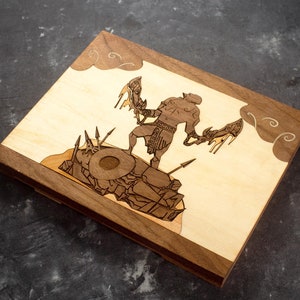 Pode incluir: Uma caixa de madeira com um design de guerreiro segurando duas espadas cortado a laser. O guerreiro est&aacute; em p&eacute; sobre uma pilha de rochas e est&aacute; rodeado de nuvens. A caixa &eacute; feita de madeira de noz e tem um acabamento castanho claro.
