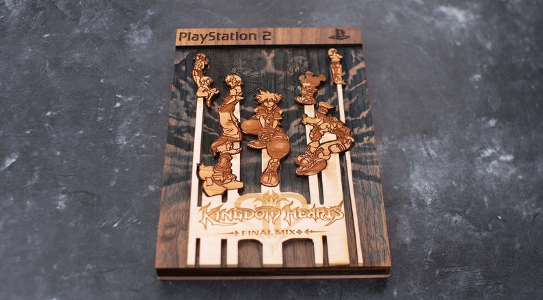 Puede incluir: Un recorte de madera de la portada del videojuego Kingdom Hearts Final Mix para Playstation 2. El recorte presenta a los personajes principales Sora, Riku y Kairi de pie en una plataforma con el t&iacute;tulo del juego y el logotipo debajo.