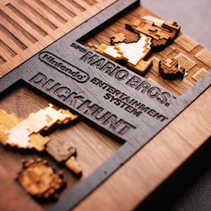 May include: A wooden replica of a Nintendo Entertainment System game cartridge featuring the Super Mario Bros. game title and Duck Hunt game title. The cartridge is made of wood and has a black border. The game characters are made of wood and are in a pixelated style.