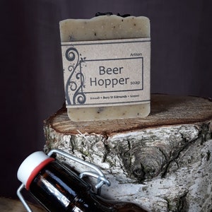 Op de afbeelding: Ambachtelijke Beer Hopper zeep met een bruin etiket met de tekst "Brewed - Bury St Edmunds - Soaked". De zeep ligt op een boomstronk met een bruine glazen fles met een metalen dop.
