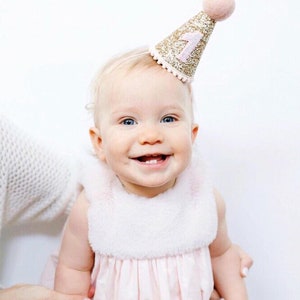 May include: A baby girl wearing a pink dress and a gold glitter party hat with a pink number one on it. The baby is smiling and looking at the camera.