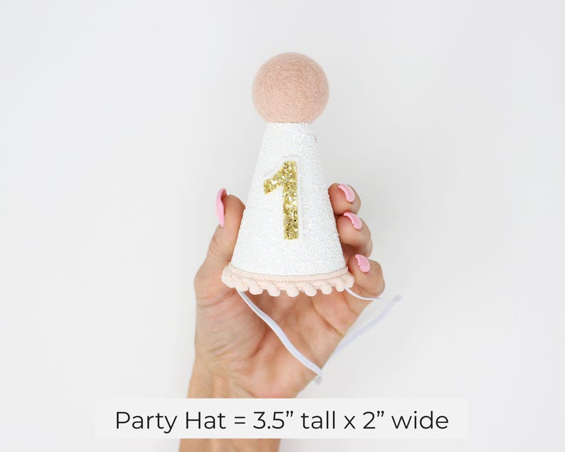 May include: White party hat with a pink pom-pom on top and a gold glitter number "1" on the front. The hat is 3.5 inches tall and 2 inches wide.
