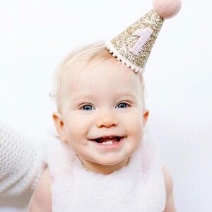 May include: A baby wearing a pink and gold glitter birthday hat with the number "1" on it. The hat has a white pom-pom on top.