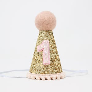 May include: Gold glitter party hat with a pink felt pom pom on top and a pink felt number one on the front. The hat has a white elastic band.