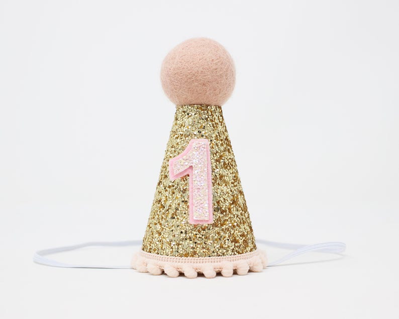 May include: Gold glitter party hat with a pink felt pom pom on top and a pink felt number one on the front. The hat has a white elastic band.