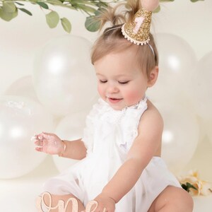 May include: A baby girl wearing a white dress and a gold glitter party hat with a pink number one on it. She is sitting in front of a white cake with a wooden 'one' topper. The cake is surrounded by white balloons.