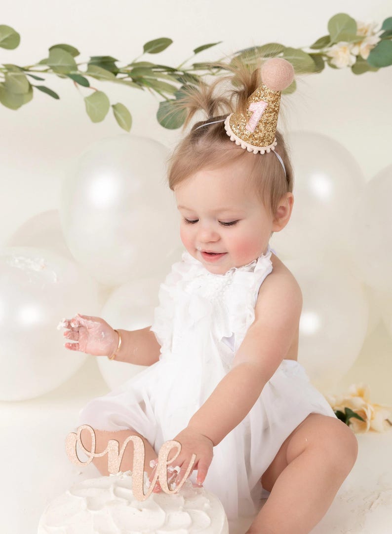 May include: A baby girl wearing a white dress and a gold glitter party hat with a pink number one on it. She is sitting in front of a white cake with a wooden 'one' topper. The cake is surrounded by white balloons.