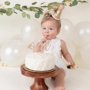 May include: A baby girl wearing a white dress and a pink and gold glitter party hat sits in front of a white cake with a gold glitter "one" topper. The cake is on a wooden cake stand. There are white balloons in the background.