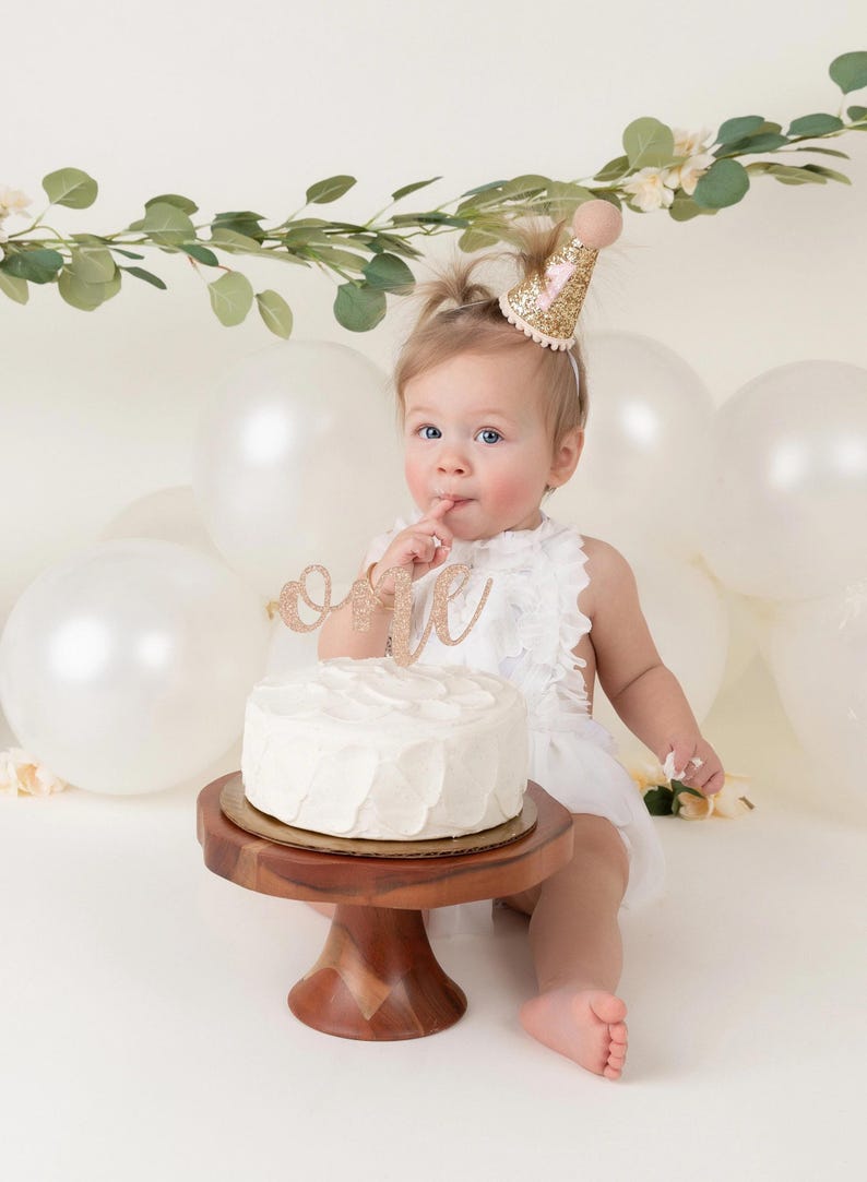 May include: A baby girl wearing a white dress and a pink and gold glitter party hat sits in front of a white cake with a gold glitter "one" topper. The cake is on a wooden cake stand. There are white balloons in the background.
