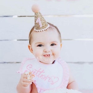 May include: A baby wearing a pink bib with the word "Sutter's" embroidered on it, is wearing a gold glitter party hat with a pink number one on it. The baby is smiling and has cake frosting on their face.