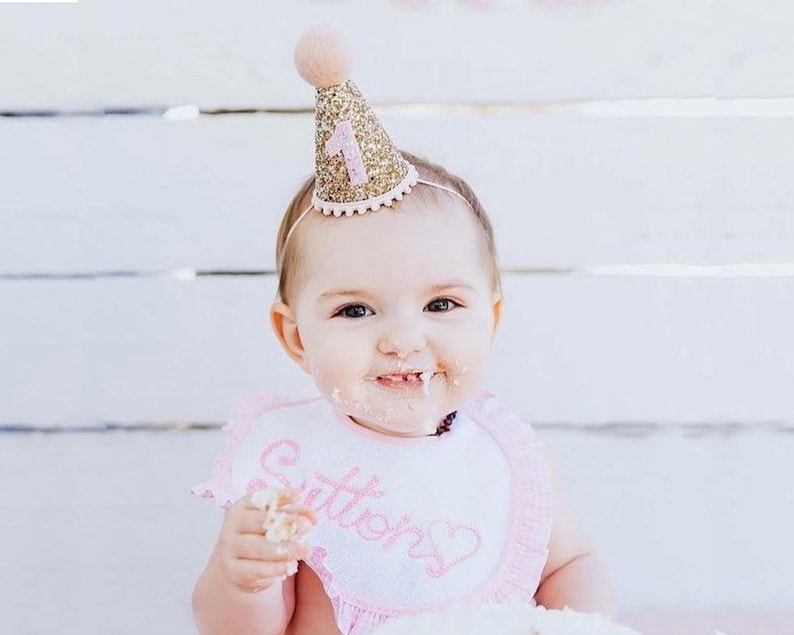May include: A baby wearing a pink bib with the word "Sutter's" embroidered on it, is wearing a gold glitter party hat with a pink number one on it. The baby is smiling and has cake frosting on their face.