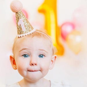 May include: A baby wearing a pink and gold glitter party hat with the number "1" on it. The hat has a pink pom-pom on top.