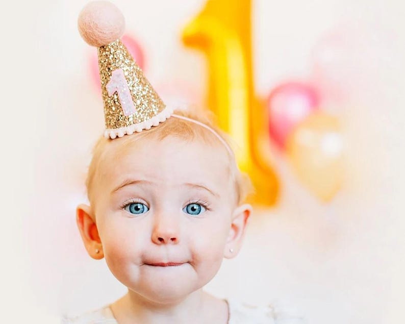 May include: A baby wearing a pink and gold glitter party hat with the number "1" on it. The hat has a pink pom-pom on top.