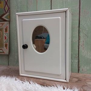 May include: White metal medicine cabinet with a round mirror and a black knob. The cabinet has a simple design and is perfect for storing toiletries or other small items.