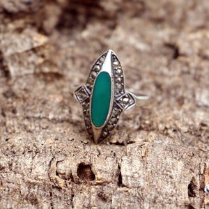 Puede incluir: Un anillo de plata con una piedra ovalada verde engastada en un diseño geométrico. El anillo está decorado con pequeñas piedras brillantes.